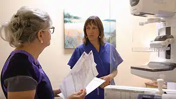 Nurse going with paper work in hand talking to patient by Mammogram machine