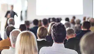 Back of peoples heads in a conference room with someone presenting up front