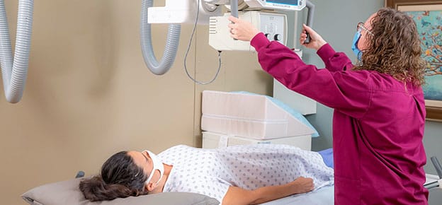 X-ray Tech adjusting the scanner over a patient. Both are wearing face masks for safety.